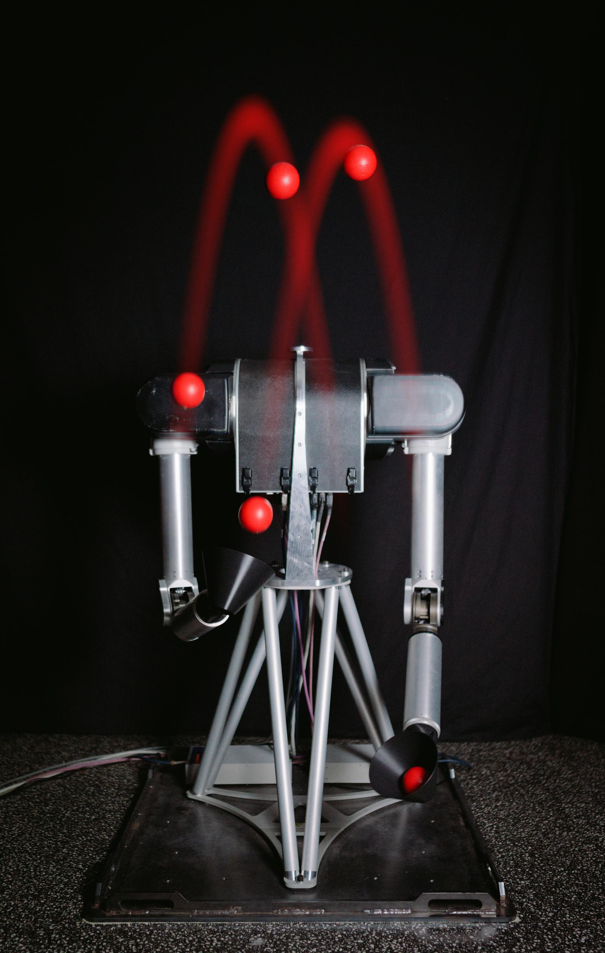 Long-exposure photograph of a Barrett WAM arm juggling, showing the ball trajectories as red arcs.
